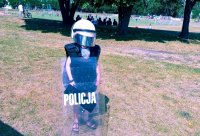 Dziecko ubrane w policyjny kask i kamizelkę, stojące z tarczą policyjną w ręku.
