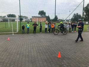Policjant obserwuje jak młody cyklista pokonuje tor przeszkód. w tle widoczna grupa uczniów przyglądająca się rowerzyście.