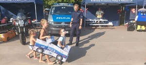 Policjant uśmiecha się do dzieci, które mają założony na siebie autochodzik imitujący policyjny radiowóz. W tle widać policyjne stoisko.