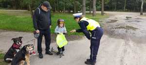 Policjantka wręczająca dziewczynce na hulajnodze odblaski. Obok dziecka stoi starszy mężczyzna z dwoma psami.