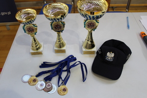Trzy puchary, medale i niebieska policyjna czapka z daszkiem leżące na białym stole.