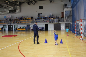 Umundurowany policjant stoi obok dziewczynki w niebieskim stroju, rozpoczynającej konkurencję sprawnościową.