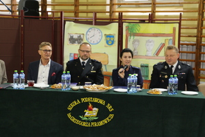Policjantka przemawia do mikrofonu, obok niej siedzi Komendant Straży Miejskiej w Pabianicach, Prezes Zarządu Ochotniczej Straży Pożarnej w Pabianicach i Starosta Pabianicki.