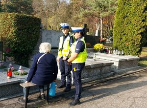 Policjantki rozmawiają ze starszą kobietą siedzącą przy grobie na cmentarzu.