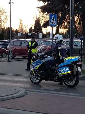 Policjant jadący na motocyklu, a obok niego stojący funkcjonariusz z tarczą do zatrzymywania pojazdów w ręku.