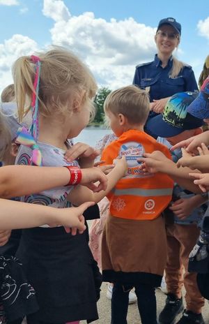 Chłopiec ma założony na plecach pomarańczowy worek promujący akcję Kręci mnie bezpieczeństwo nad wodą. Dzieci palcami pokazują na niego. W tle widać uśmiechającą się policjantkę.