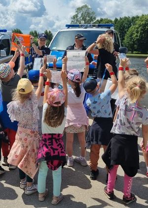 Policjantka pokazuje dzieciom pomarańczowy worek, a dzieci mają wyciągnięte do góry ręce, w których trzymają upominki. Obok funkcjonariuszki stoi strażnik miejski, a za nią widoczny jest policyjny radiowóz.