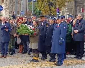 Delegacja służb mundurowych, przed nią stoi harcerka trzymająca wiązankę kwiatów.