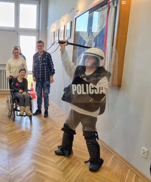 Uczestnik Warsztatów Terapii Zajęciowej pozuje ubrany w policyjny sprzęt. W tle widać funkcjonariusza i osobę na wózku inwalidzkim.