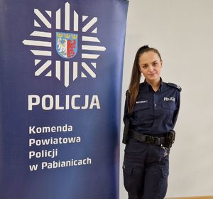Policjantka stoi przy banerze z logo pabianickiej Policji oraz z napisem Komenda Powiatowa Policji w Pabianicach.