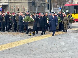 Uczestnicy obchodów idą pod Pomnik Legionisty. Na ich czele widać młodą harcerkę niosącą wiązankę kwiatów.