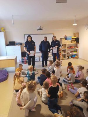 Policjantka i dwie strażniczki miejskie stoją przodem przed grupą dzieci, trzymają dyplomy.