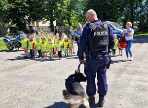 Na pierwszym planie przewodnik psa służbowego ze swoim kompanem. Na drugim planie grupa przedszkolaków w kamizelkach odblaskowych.