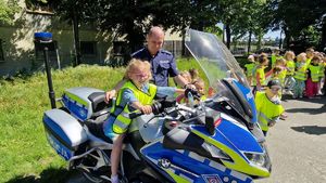 Dziecko siedzi na motocyklu, obok stoi policjant. Na drugim planie  znajduje się grupa przedszkolaków w kamizelkach odblaskowych.