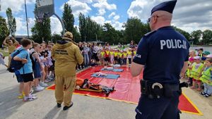 Policjant i strażak stoją na pierwszym planie, dalej widać rozłożoną czerwoną plandekę na której znajdują się fantomy. Wokół plandeki stoją dzieci.