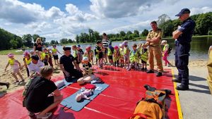 Na czerwonej plandece są rozłożone fantomy. Policjant klęczy obok i pokazuje, zgromadzonym widzom, pierwszą pomoc przedmedyczną.