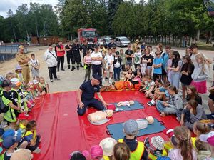 Na czerwonej plandece są rozłożone fantomy. Policjant klęczy obok i pokazuje, zgromadzonym widzom, pierwszą pomoc przedmedyczną