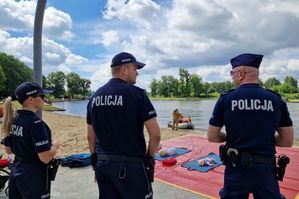 Policjanci stoją na tle czerwonej plandeki i zbiornika wodnego.