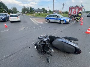 Miejsce wypadku drogowego. Na pierwszym planie widać leżący motocykl, w tle stoi policyjnym radiowóz, obok przejeżdżają auta osobowe.