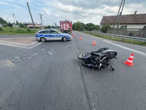 Miejsce wypadku drogowego. Na pierwszym planie widać leżący motocykl, w tle stoi policyjnym radiowóz, obok przejeżdżają auta osobowe.