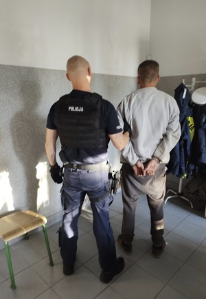 Policjant i mężczyzna stoją tyłem. Policjant trzyma mężczyznę za rękę. Mężczyzna ma założone kajdanki na ręce trzymane z tyłu.