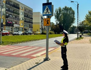 Policjantka stoi przy przejściu dla pieszych i patrzy na znaki drogowe. W tle widać bloki mieszkalne i pojazdy.