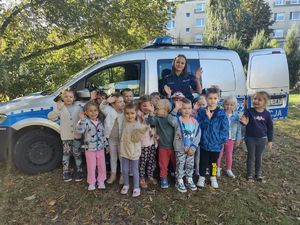 Policjantka stoi z dziećmi na tle oznakowanego radiowozu.