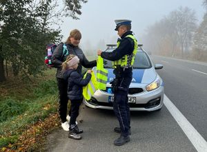 Policjant wręcza dziewczynce kamizelkę odblaskową, obok stoi kobieta i się uśmiecha.