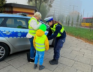 Policjantka zakłada dziecku kamizelkę odblaskową. Obok stoi kobieta i ubiera kamizelkę odblaskową.
