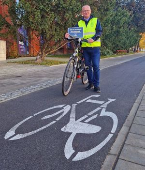 Starosta pabianicki stoi na ścieżce rowerowej i trzyma w ręku czarną kartkę z napisem #dajsięzauważyć #nośodblaski# .