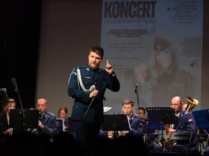 Dyrygent wskazujący palcem w górę. Za policjantem widać pozostałych muzyków, a w tle wyświetlony jest plakat wydarzenia.