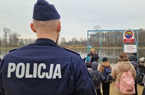 Na pierwszym planie widać policjanta. Dalej, na tle zbiornika wodnego strażak rozmawia z dziećmi.