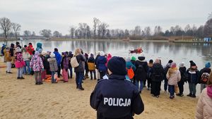 Policjantka stoi na brzegu i obserwuje dzieci patrzące jak strażak wyciąga z wody poszkodowanego.