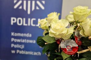 Białe róże na tle baneru z napisem Komenda Powiatowa Policji w Pabianicach.