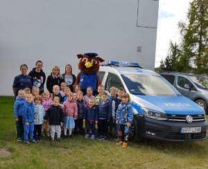 Dzieci wraz z policjantką i maskotką policji stoją na tle radiowozu policyjnego.