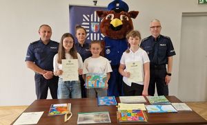 Policjanci pozują do wspólnego zdjęcia z laureatami konkursu. Przed nimi znajdują się wszystkie prace konkursowe.