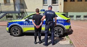 Na tle radiowozu policjant trzyma osobę zatrzymaną.