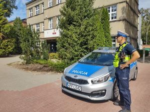 Umundurowany policjant stoi przed oznakowanym radiowozem. W tle szkoła.