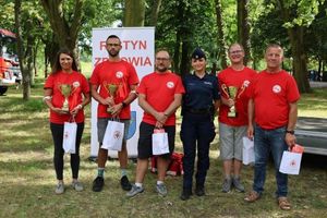 Policjantka stoi wraz z uczestnikami turnieju wiedzy. Uczestnicy ubrani w czerwone koszulki Polskiego Czerwonego Krzyża.