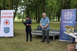 Policjantka wraz z mężczyzną - prezesem Polskiego Czerwonego Krzyża stoją przed sceną. W tle dwa banery z napisami policja oraz festyn rodzinny.