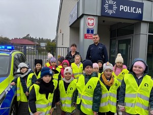 Dzieci w kamizelkach odblaskowych stoją wraz z nauczycielką i policjantem przed budynkiem Posterunku Policji we Wrzącej.