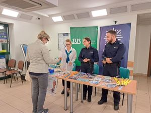 Policjanci i pracownica miejscowego Zakładu Ubezpieczeń Społecznych stoją przy stoisku profilaktycznym, przy którym znajduje się również zainteresowana inicjatywą kobieta.