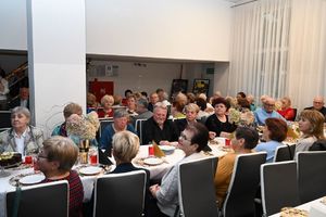 Zgromadzeni na spotkaniu seniorzy siedzący przy zastawionych stołach.