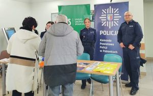 Policjant, policjantka i pracownica Zakładu Ubezpieczeń Społecznych przy stoisku informacyjnym. Na przeciwko nich stoją seniorzy.