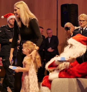 Dziewczynka odchodzi od Świętego Mikołaja, jest prowadzona przez kobietę a w rękach trzyma czekoladki.