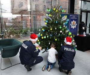 Dziewczynka w towarzystwie dwóch mundurowych ubiera odblaskową choinkę.