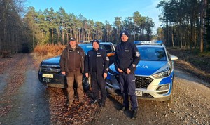 Policjanci razem z Komendantem Posterunku Straży Leśnej stoją w lesie. Za funkcjonariuszami auta służbowe.