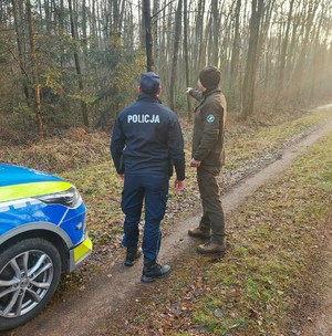 Policjant i strażnik w lesie, stoją przy oznakowanym radiowozie.