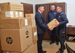 Mężczyzna przekazuje zapakowane pudło policjantowi. Obok stoi drugi policjant. Obok kilka innych zapakowanych kartonów.