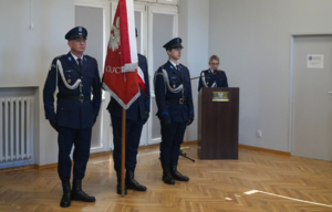 Lektor czyta przy mównicy treść scenariusza, obok stoi poczet sztandarowy Komendy Powiatowej Policji w Pabianicach.